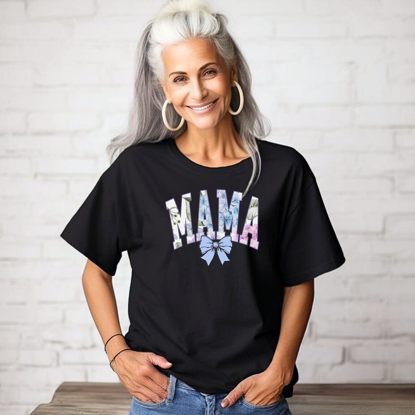 Person wearing a black T-shirt featuring the word “MAMA” in large block letters filled with a delicate blue hydrangea floral pattern, accented with soft green and pink tones. Below the text is a pastel blue coquette bow, adding a sweet, feminine touch. This spring-inspired graphic tee blends floral elegance with playful charm, perfect for fashionable moms.