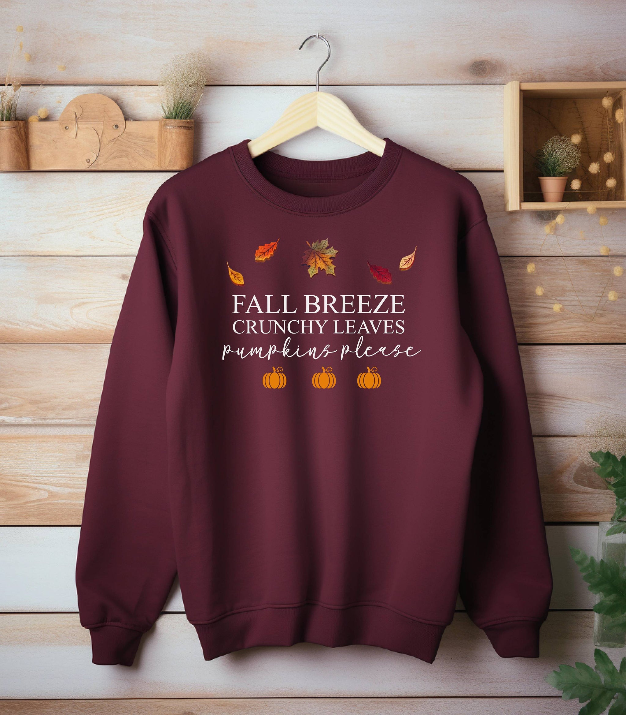 Maroon Gildan sweatshirt featuring a festive fall-themed design with colorful autumn leaves, the phrase “Fall Breeze, Crunchy Leaves, Pumpkins Please” in mixed fonts, and four orange pumpkin icons along the bottom. A cozy and stylish piece perfect for embracing crisp air, seasonal vibes, and pumpkin spice everything.