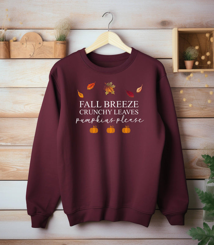 Maroon Gildan sweatshirt featuring a festive fall-themed design with colorful autumn leaves, the phrase “Fall Breeze, Crunchy Leaves, Pumpkins Please” in mixed fonts, and four orange pumpkin icons along the bottom. A cozy and stylish piece perfect for embracing crisp air, seasonal vibes, and pumpkin spice everything.