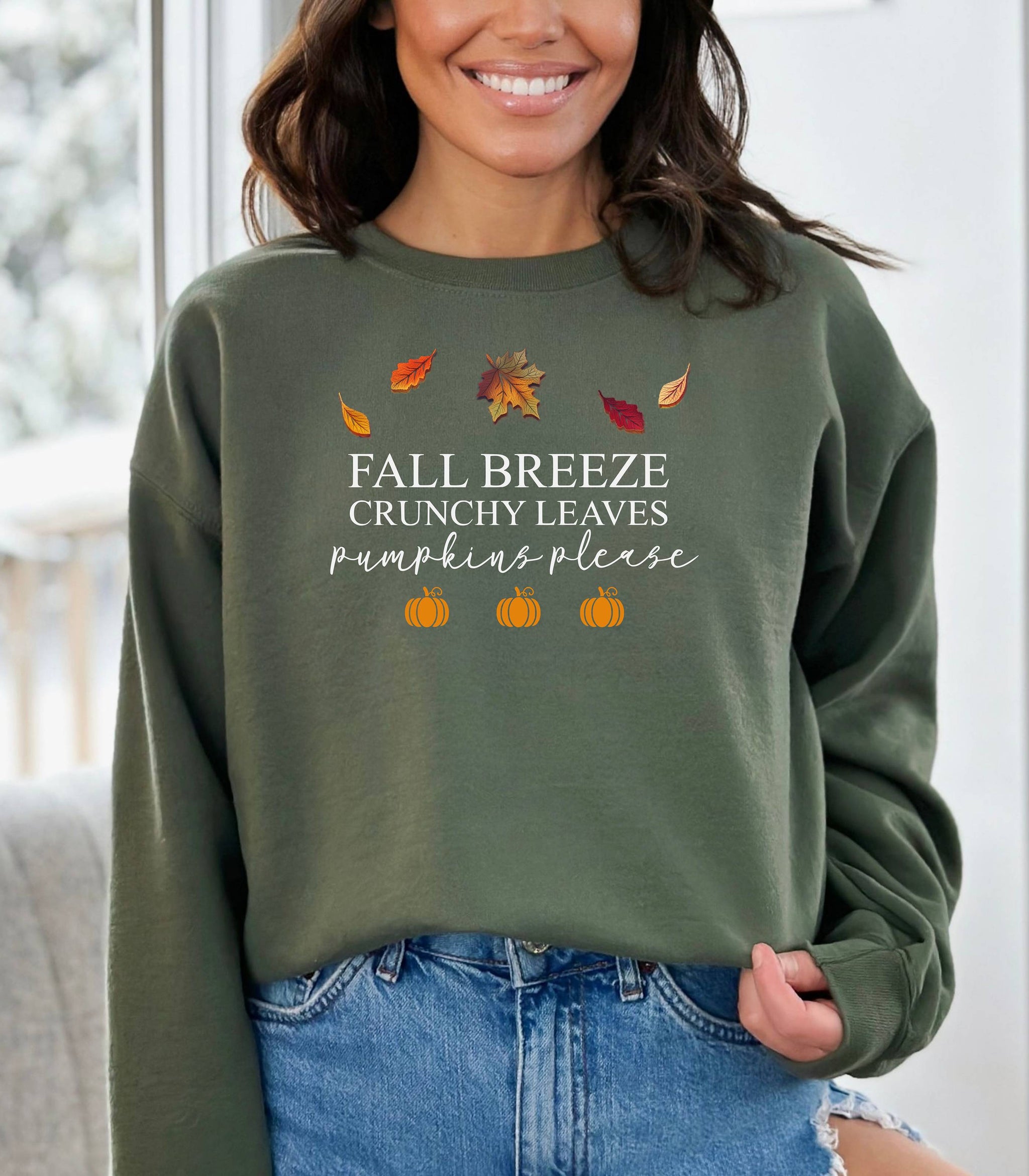 Military Green Gildan sweatshirt featuring a festive fall-themed design with colorful autumn leaves, the phrase “Fall Breeze, Crunchy Leaves, Pumpkins Please” in mixed fonts, and four orange pumpkin icons along the bottom. A cozy and stylish piece perfect for embracing crisp air, seasonal vibes, and pumpkin spice everything.
