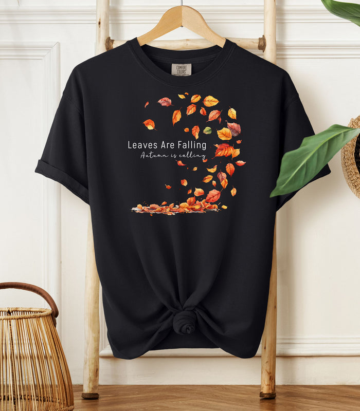 Black Comfort Colors T-shirt featuring a fall-themed design with swirling autumn leaves and the phrase "Leaves Are Falling, Autumn is Calling" in elegant white script. The vibrant orange, red, and yellow leaves create a whimsical motion effect. Shirt is styled with a front knot and displayed on a wooden hanger in a cozy, natural setting.
