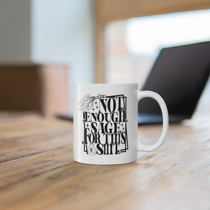 White ceramic mug featuring bold black typography reading “Not Enough Sage For This Shit,” framed in a whimsical outlined border. The design includes a detailed sage bundle illustration at the top and a small jar with a sage leaf motif at the bottom, adding a witchy, mystical vibe. Perfect for humorous coffee or tea moments, it blends sassy attitude with boho and spiritual flair, making it an ideal gift for friends with a sense of humor.