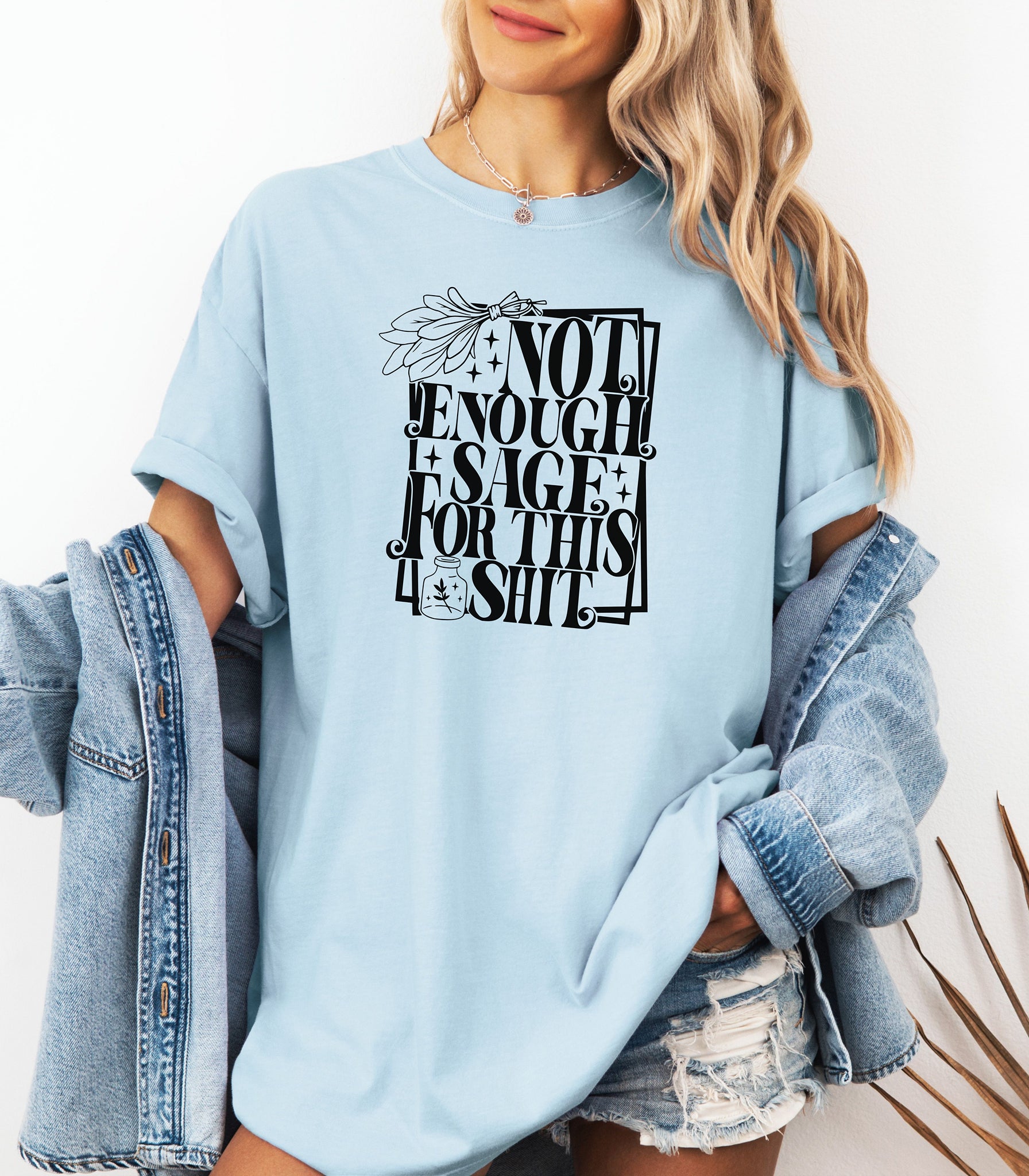 Chambray light blue T-shirt with bold black typography reading “Not Enough Sage For This Shit,” accented with illustrated sage leaves, sparkles, and a small potion jar with a leafy design. Playful and cheeky statement shirt for spiritual, witchy, or humor-loving personalities.