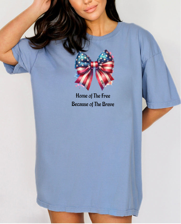 Washed Denim t-shirt featuring a patriotic coquette bow design with stars and stripes in red, white, and blue. Below the bow, the text reads 'Home of The Free Because of The Brave.' A stylish and feminine Holiday shirt, perfect for celebrating Independence Day, parades, and summer festivities.