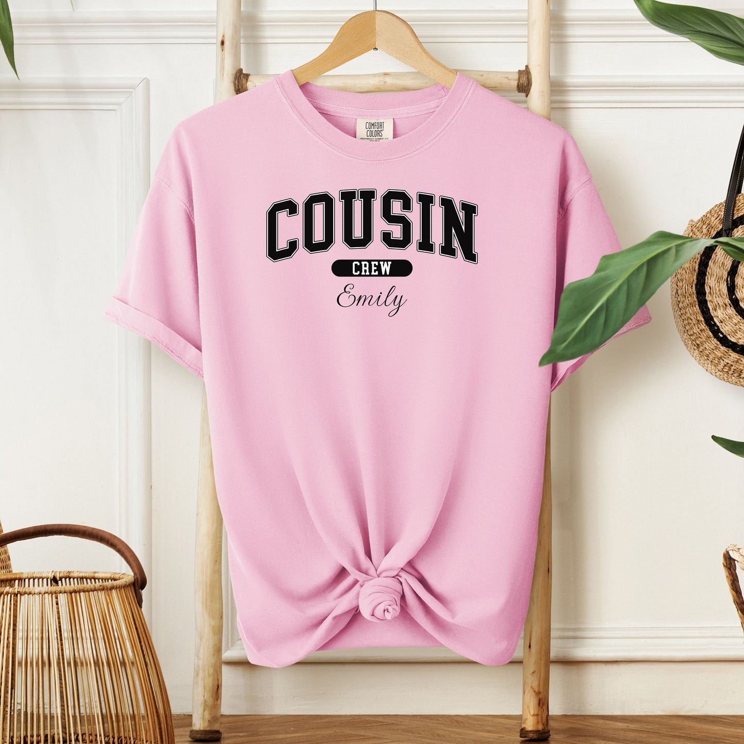 Blossom pink Comfort Colors T-shirt featuring a bold “Cousin Crew” design in black varsity-style lettering, with the name “Your Name” in elegant script below. Displayed knotted at the bottom and hanging on a wooden hanger in a cozy, neutral-toned room with natural decor elements.