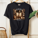 A black t-shirt featuring a whimsical "Read More Books, Magick Awaits" design. The graphic showcases an open enchanted book surrounded by candles, a crescent moon, owl, pocket watch, hourglass, and magical elements, creating a mystical and literary aesthetic perfect for book lovers and fantasy fans.