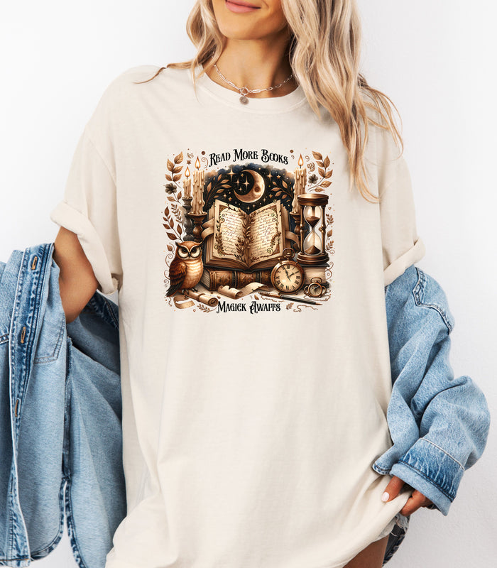 Person wearing an ivory t-shirt featuring a whimsical "Read More Books, Magick Awaits" design. The graphic showcases an open enchanted book surrounded by candles, a crescent moon, owl, pocket watch, hourglass, and magical elements, creating a mystical and literary aesthetic perfect for book lovers and fantasy fans.