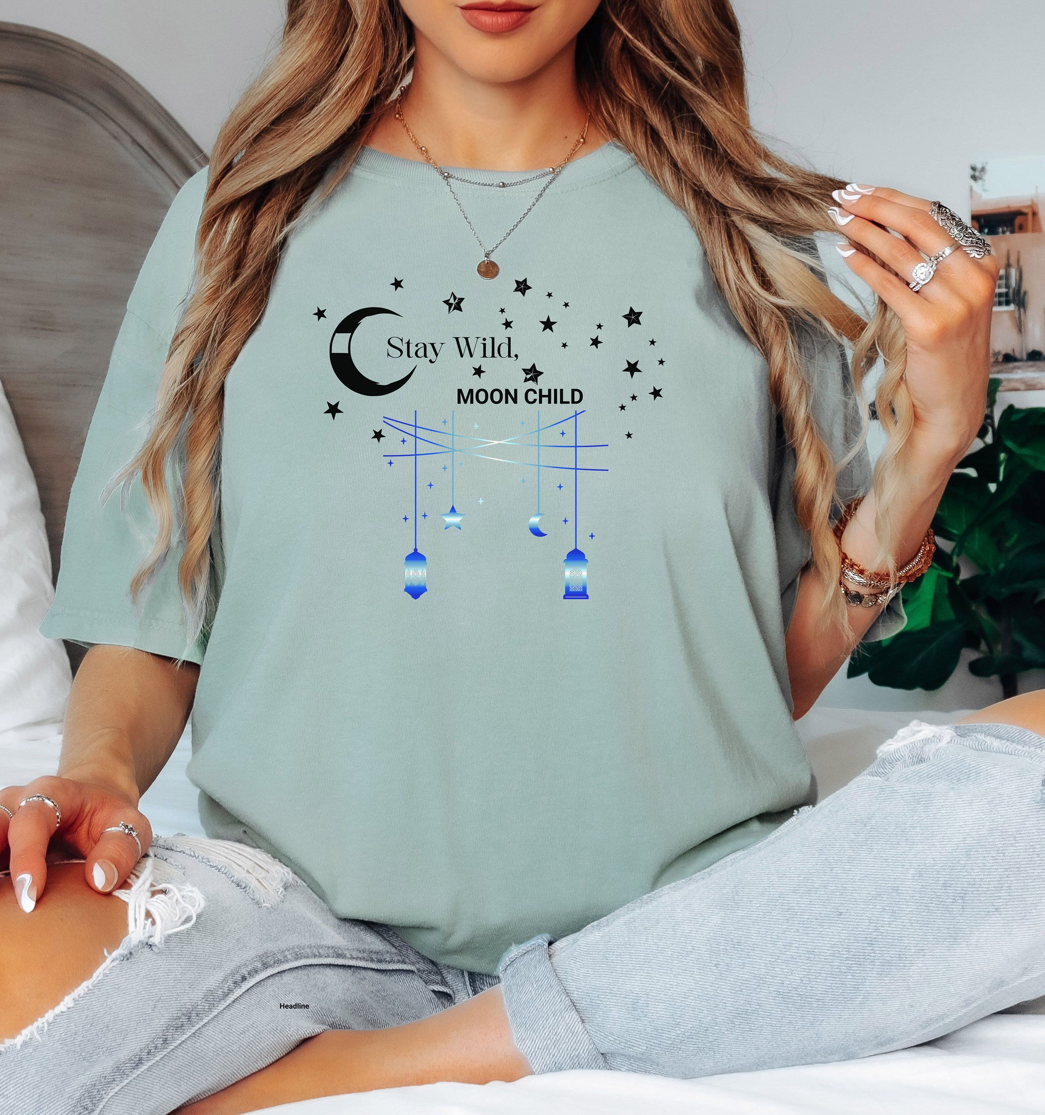 Stay Wild Moon Child T-shirt featuring a celestial design with a crescent moon, stars, and hanging lanterns. The text 'Stay Wild, Moon Child' is surrounded by cosmic elements, evoking a dreamy bohemian vibe. Printed on a bay unisex tee, this mystical and free-spirited graphic is perfect for astrology lovers, night sky dreamers, and those who embrace their wild, cosmic side.