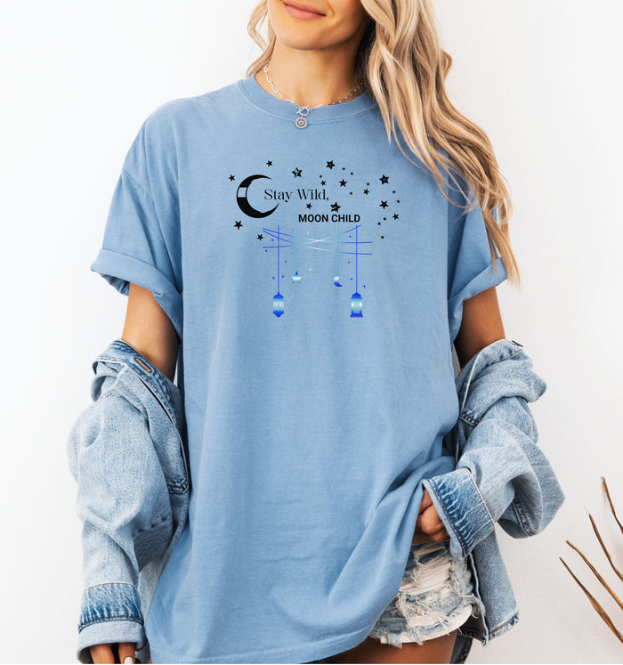 Stay Wild Moon Child T-shirt featuring a celestial design with a crescent moon, stars, and hanging lanterns. The text 'Stay Wild, Moon Child' is surrounded by cosmic elements, evoking a dreamy bohemian vibe. Printed on a washed denim unisex tee, this mystical and free-spirited graphic is perfect for astrology lovers, night sky dreamers, and those who embrace their wild, cosmic side.