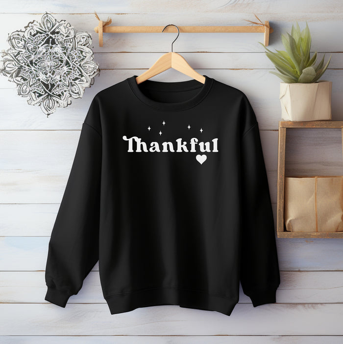 Black Gildan sweatshirt featuring the word “Thankful” in bold white retro-style font, accented with small sparkling stars above and a heart below. A simple yet meaningful design that celebrates gratitude and cozy fall vibes—perfect for Thanksgiving, autumn gatherings, or everyday wear.