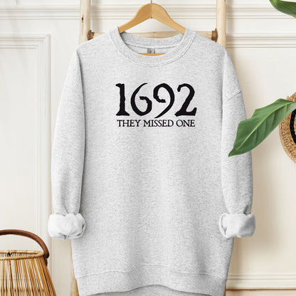 Ash sweatshirt with '1692 THEY MISSED ONE' text on a hanger against a white wall.