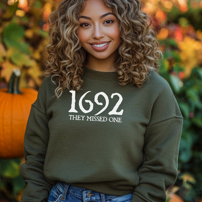 Person wearing a military green sweatshirt with '1692 THEY MISSED ONE' text, standing in front of pumpkins and autumn foliage.