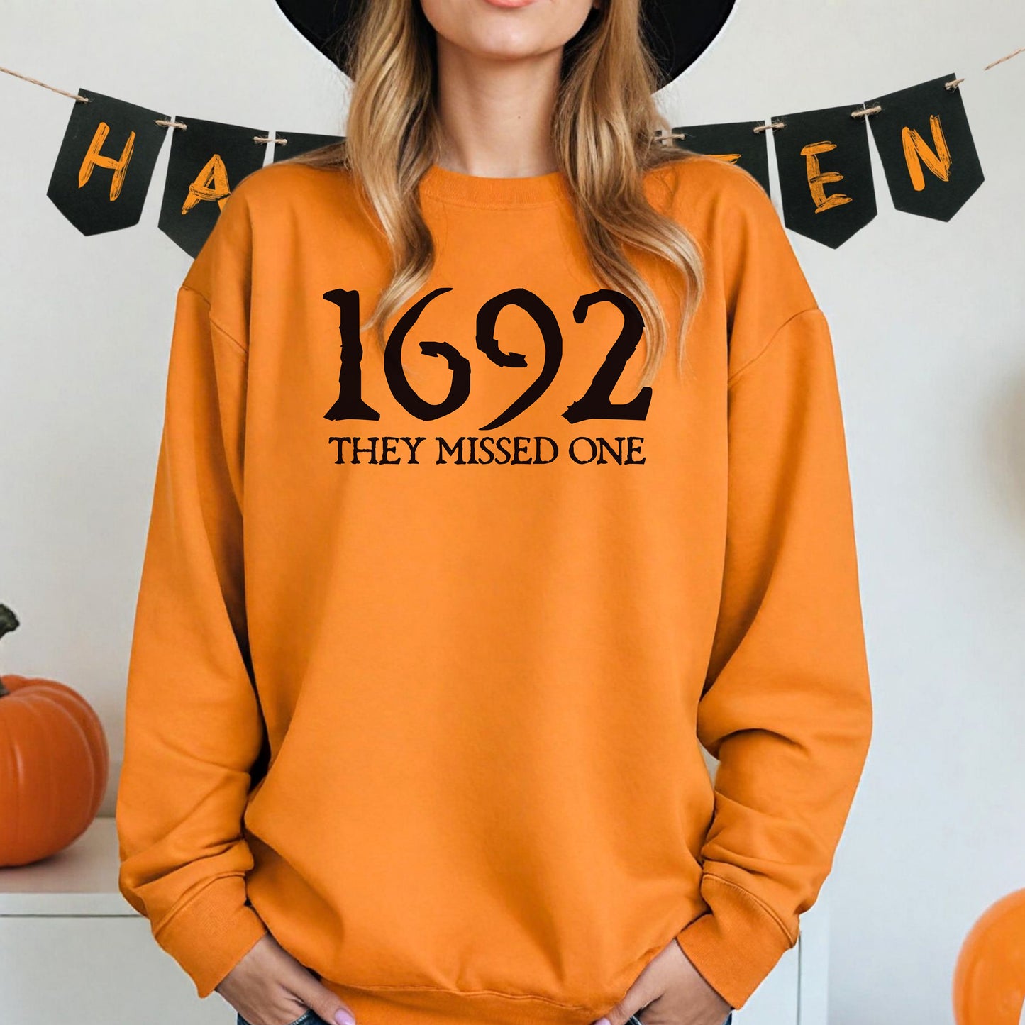 Person wearing a safety orange sweatshirt with '1692 THEY MISSED ONE' text, standing in front of Halloween-themed decorations.
