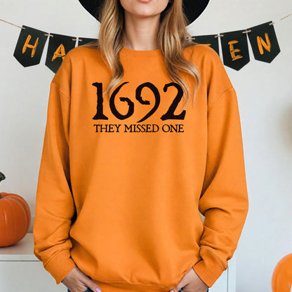 Person wearing a safety orange sweatshirt with '1692 THEY MISSED ONE' text, standing in front of Halloween-themed decorations.