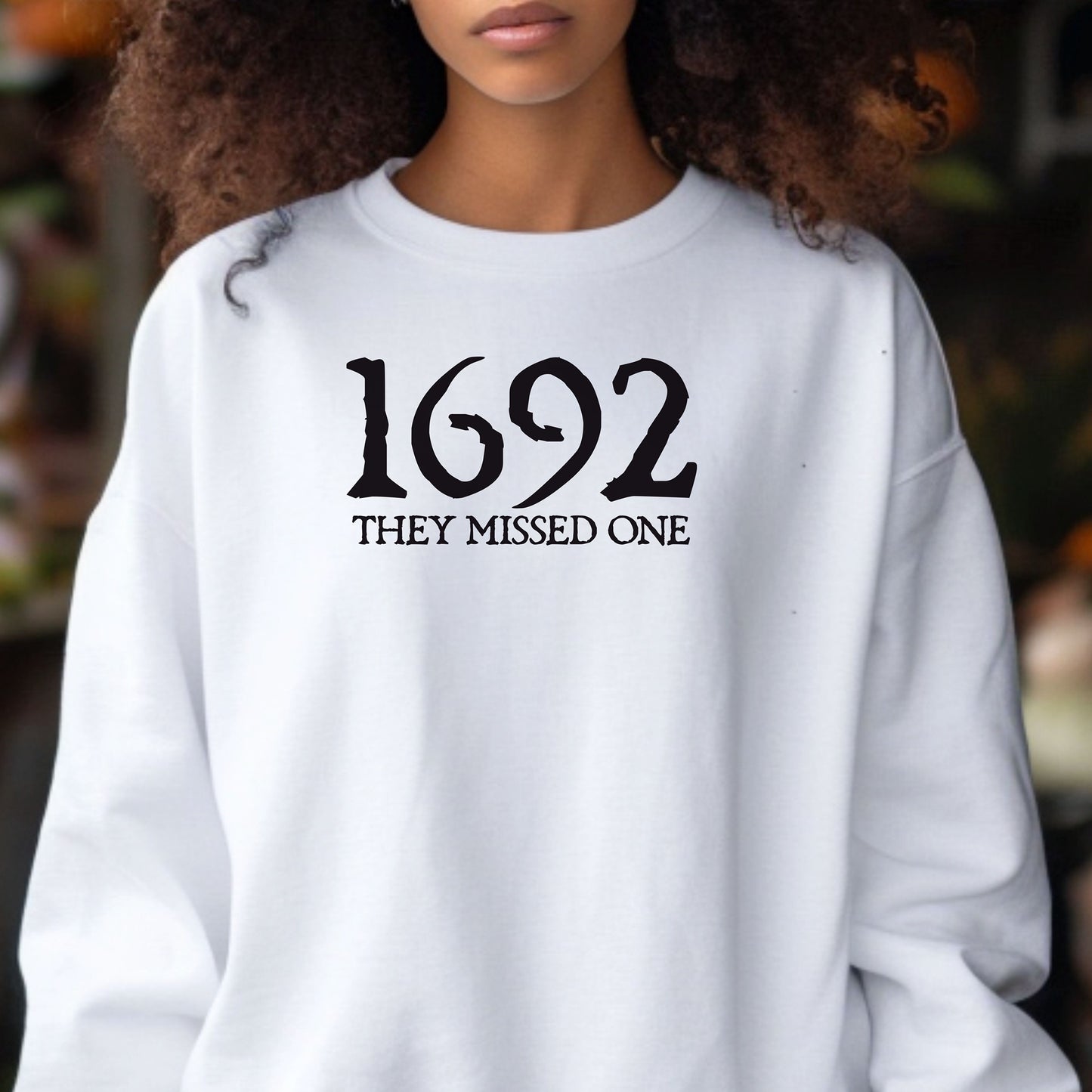 Person wearing an Ash sweatshirt with '1692 THEY MISSED ONE' text outdoor in an autumn setting.