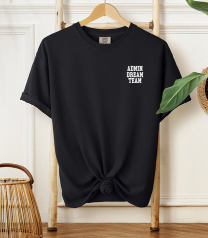 Black Comfort Colors t-shirt hanging on a wooden hanger, featuring a minimalist white text design on the left chest that reads "ADMIN DREAM TEAM." Perfect for school staff, office teams, or administrative professionals showing team spirit in a stylish and understated way.