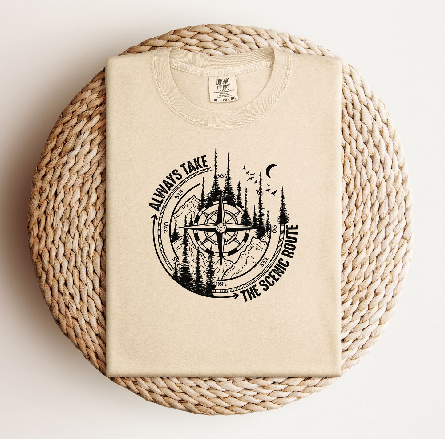 Always Take The Scenic Route T-shirt featuring a detailed compass design surrounded by tall pine trees, mountains, a crescent moon, and flying birds. This outdoor adventure graphic symbolizes exploration, travel, and a love for nature. Printed on a soft, comfortable ivory unisex tee, it’s perfect for hikers, campers, and wanderers who embrace the beauty of the journey and prefer the scenic path over the fast route.