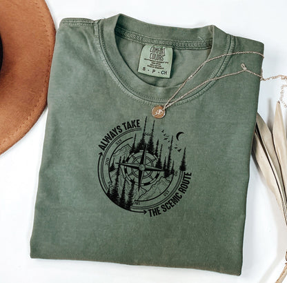 Always Take The Scenic Route T-shirt featuring a detailed compass design surrounded by tall pine trees, mountains, a crescent moon, and flying birds. This outdoor adventure graphic symbolizes exploration, travel, and a love for nature. Printed on a soft, comfortable moss unisex tee, it’s perfect for hikers, campers, and wanderers who embrace the beauty of the journey and prefer the scenic path over the fast route.