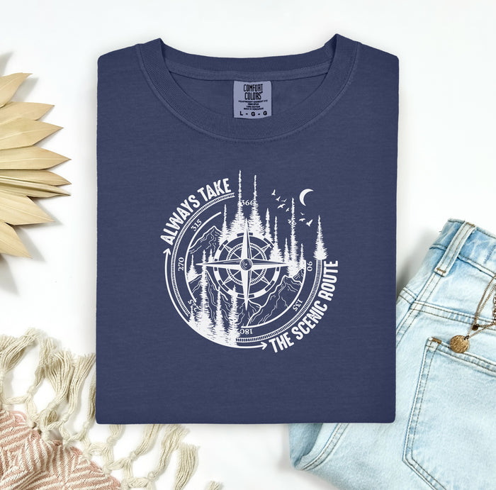 Always Take The Scenic Route T-shirt featuring a detailed compass design surrounded by tall pine trees, mountains, a crescent moon, and flying birds. This outdoor adventure graphic symbolizes exploration, travel, and a love for nature. Printed on a soft, comfortable navy unisex tee, it’s perfect for hikers, campers, and wanderers who embrace the beauty of the journey and prefer the scenic path over the fast route.