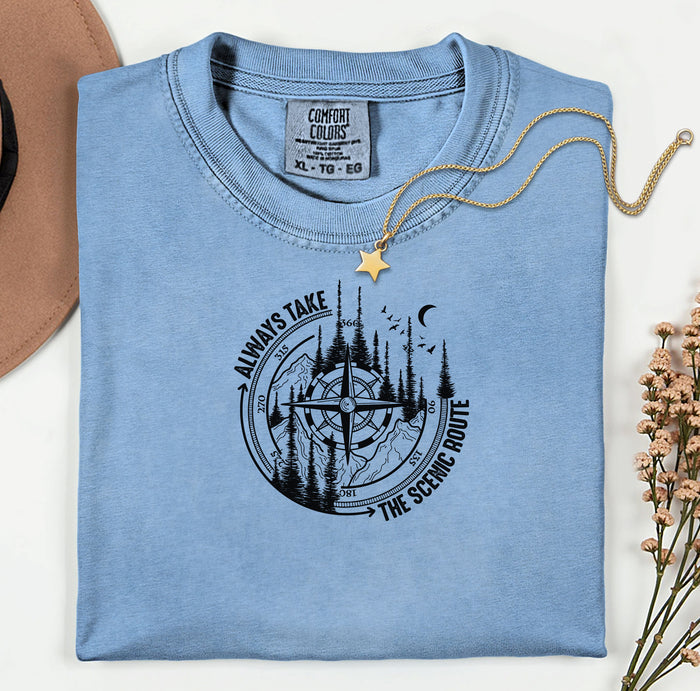 Always Take The Scenic Route T-shirt featuring a detailed compass design surrounded by tall pine trees, mountains, a crescent moon, and flying birds. This outdoor adventure graphic symbolizes exploration, travel, and a love for nature. Printed on a soft, comfortable washed denim unisex tee, it’s perfect for hikers, campers, and wanderers who embrace the beauty of the journey and prefer the scenic path over the fast route.