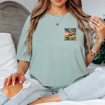 Bay light sage t-shirt featuring a patriotic pocket design with an American flag waving over a scenic farmland landscape at sunset. The design includes the text 'USA EST 1776,' symbolizing pride and heritage. Perfect for 4th of July, country living, and casual patriotic style with a subtle, classic look.