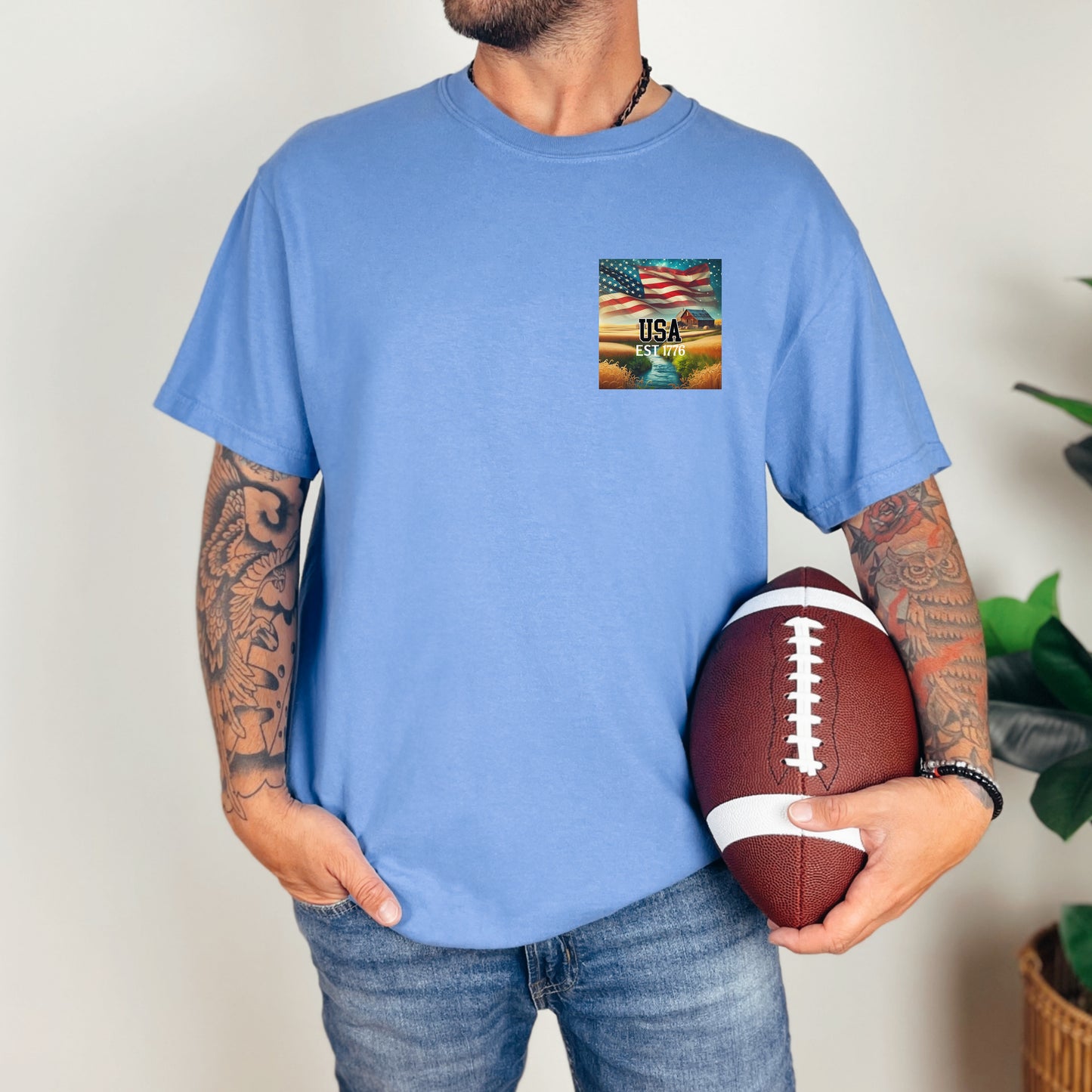 Mystic Blue t-shirt featuring a patriotic pocket design with an American flag waving over a scenic farmland landscape at sunset. The design includes the text 'USA EST 1776,' symbolizing pride and heritage. Perfect for 4th of July, country living, and casual patriotic style with a subtle, classic look.
