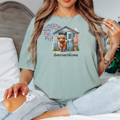 Bay light sage green t-shirt featuring a 'Barnyard Freedom' design with a cute Highland cow standing in front of a rustic blue barn decorated with an American flag. Red, white, and blue fireworks burst in the background, creating a festive patriotic country vibe. Perfect for 4th of July and farm life lovers.