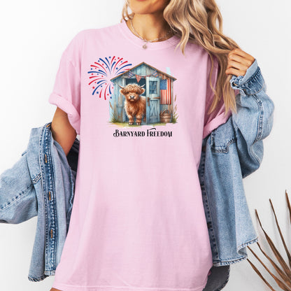 Blossom pink t-shirt featuring a 'Barnyard Freedom' design with a cute Highland cow standing in front of a rustic blue barn decorated with an American flag. Red, white, and blue fireworks burst in the background, creating a festive patriotic country vibe. Perfect for 4th of July and farm life lovers.