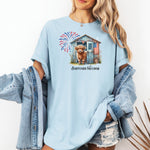 Chambray light blue t-shirt featuring a 'Barnyard Freedom' design with a cute Highland cow standing in front of a rustic blue barn decorated with an American flag. Red, white, and blue fireworks burst in the background, creating a festive patriotic country vibe. Perfect for 4th of July and farm life lovers.
