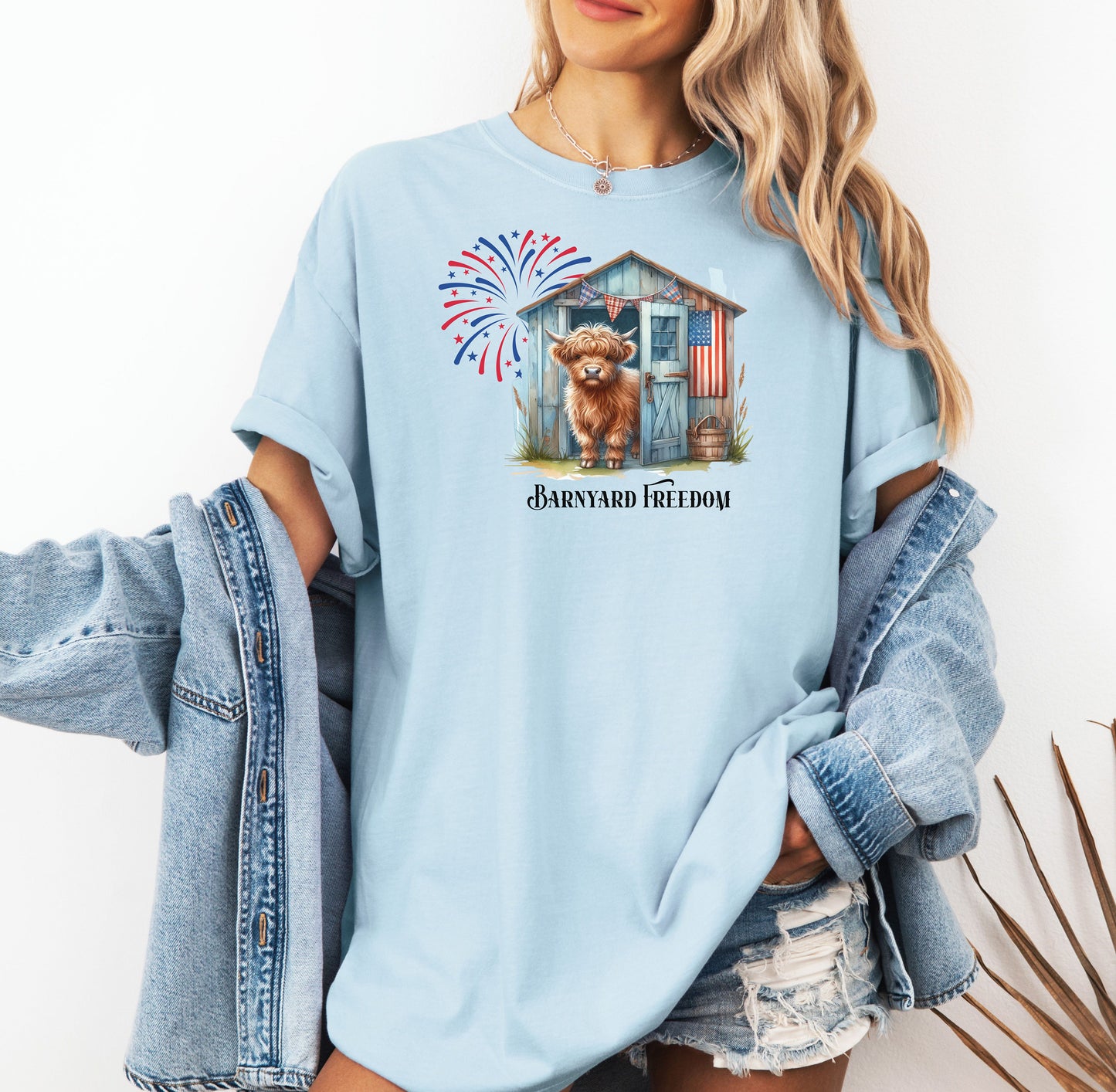 Chambray light blue t-shirt featuring a 'Barnyard Freedom' design with a cute Highland cow standing in front of a rustic blue barn decorated with an American flag. Red, white, and blue fireworks burst in the background, creating a festive patriotic country vibe. Perfect for 4th of July and farm life lovers.