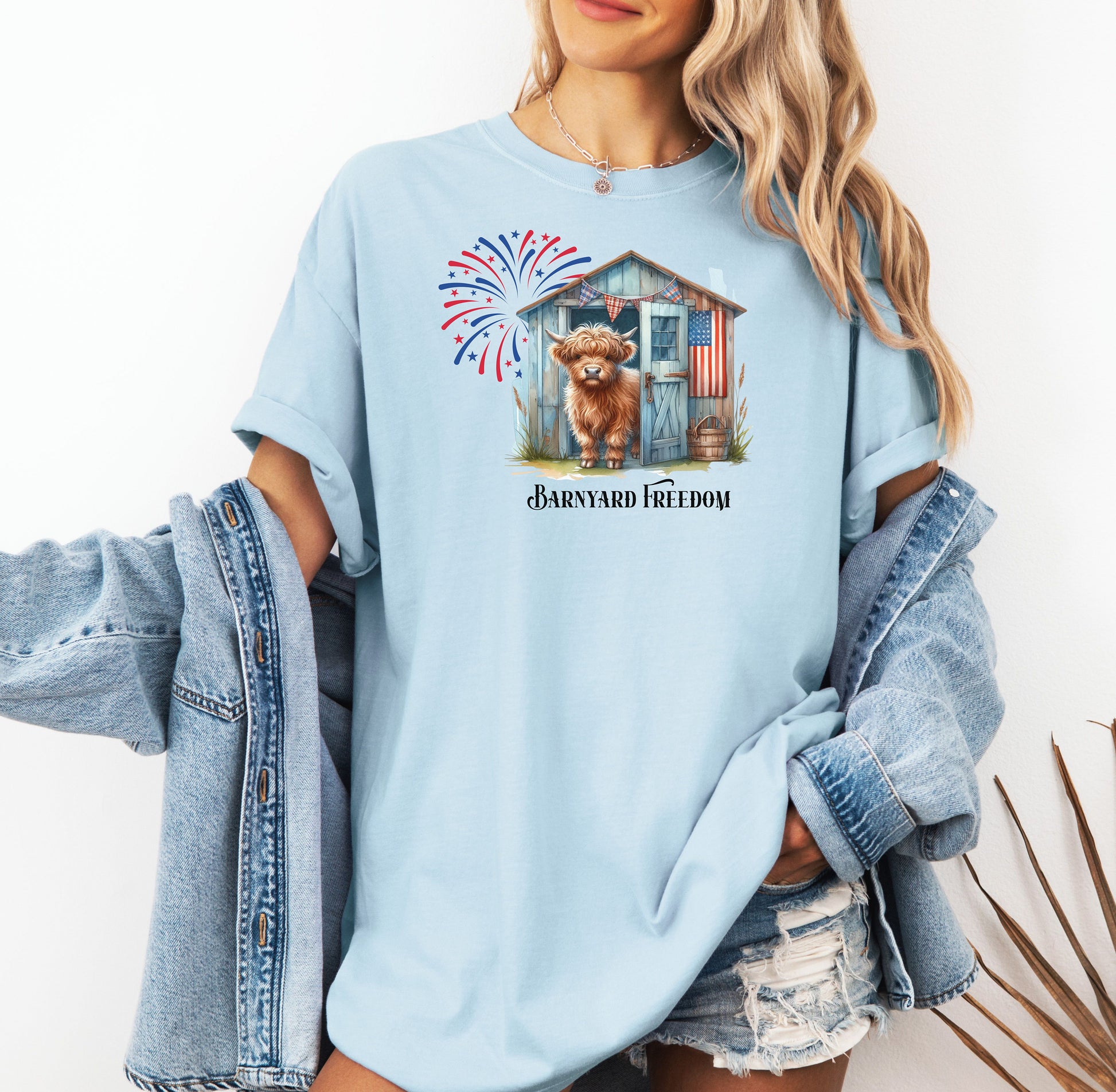 Chambray light blue t-shirt featuring a 'Barnyard Freedom' design with a cute Highland cow standing in front of a rustic blue barn decorated with an American flag. Red, white, and blue fireworks burst in the background, creating a festive patriotic country vibe. Perfect for 4th of July and farm life lovers.