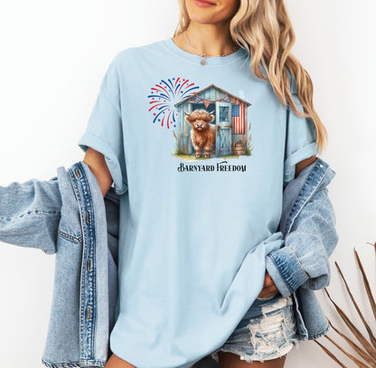 Chambray light blue t-shirt featuring a 'Barnyard Freedom' design with a cute Highland cow standing in front of a rustic blue barn decorated with an American flag. Red, white, and blue fireworks burst in the background, creating a festive patriotic country vibe. Perfect for 4th of July and farm life lovers.