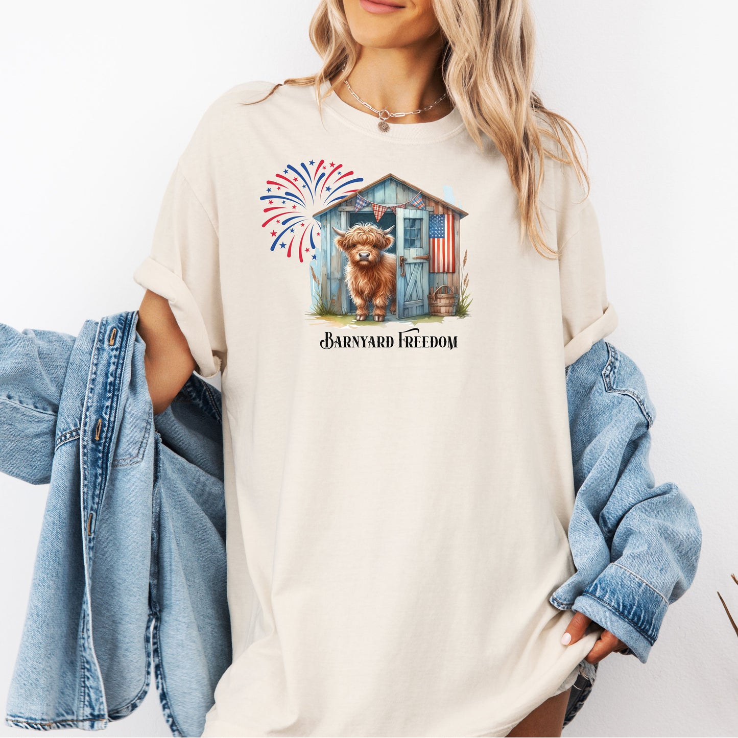 Ivory t-shirt featuring a 'Barnyard Freedom' design with a cute Highland cow standing in front of a rustic blue barn decorated with an American flag. Red, white, and blue fireworks burst in the background, creating a festive patriotic country vibe. Perfect for 4th of July and farm life lovers.
