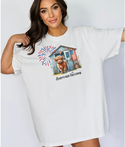 White t-shirt featuring a 'Barnyard Freedom' design with a cute Highland cow standing in front of a rustic blue barn decorated with an American flag. Red, white, and blue fireworks burst in the background, creating a festive patriotic country vibe. Perfect for 4th of July and farm life lovers.