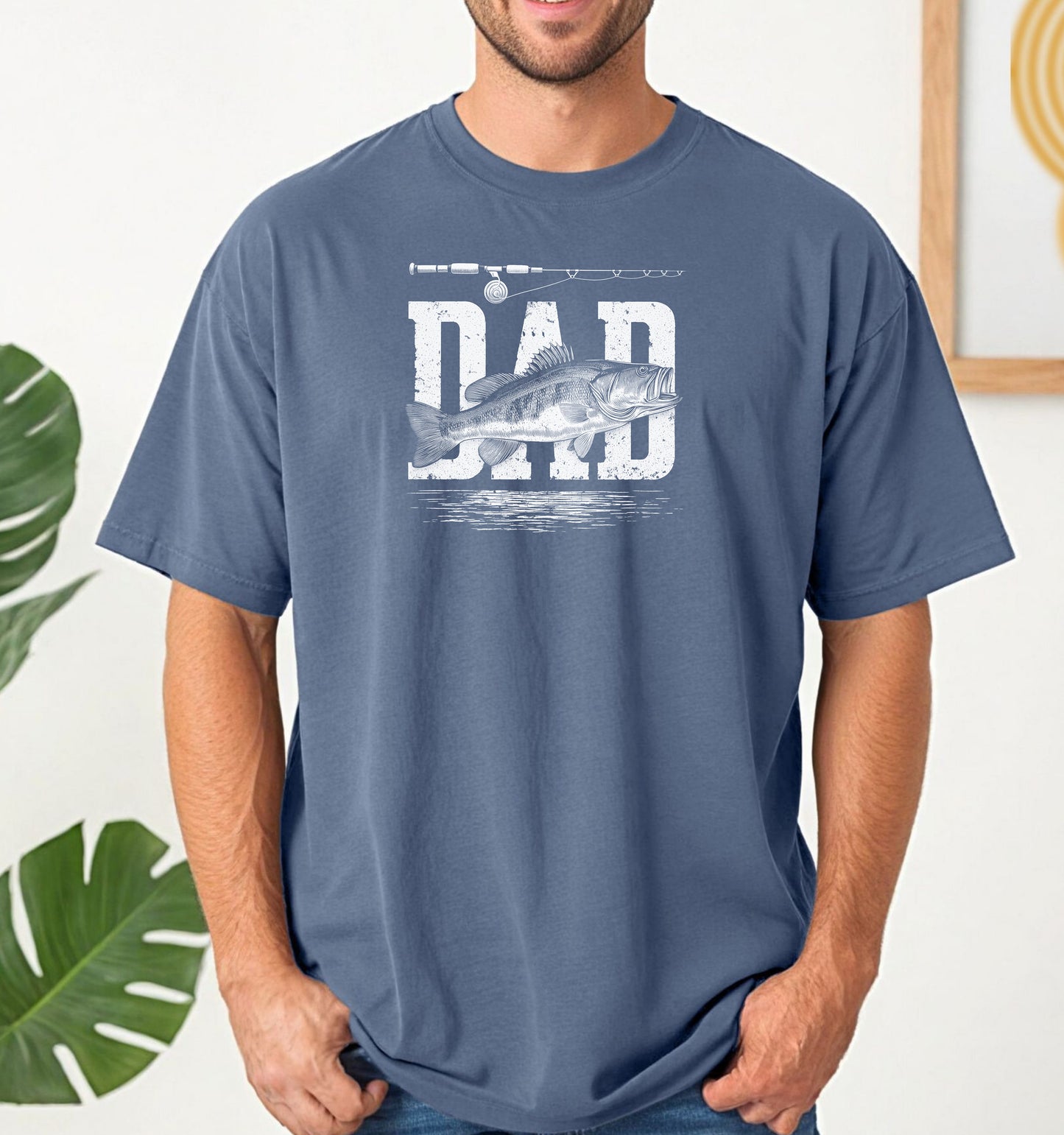 Bass Fishing Dad T-shirt featuring the word 'DAD' in bold, distressed white lettering with a detailed bass fish illustration overlaying the text. A fishing rod graphic runs above the design, with subtle water ripple details below. Printed on a blue jean unisex tee, this shirt is perfect for dads who love fishing, making it an ideal gift for Father’s Day, birthdays, or weekend fishing trips.