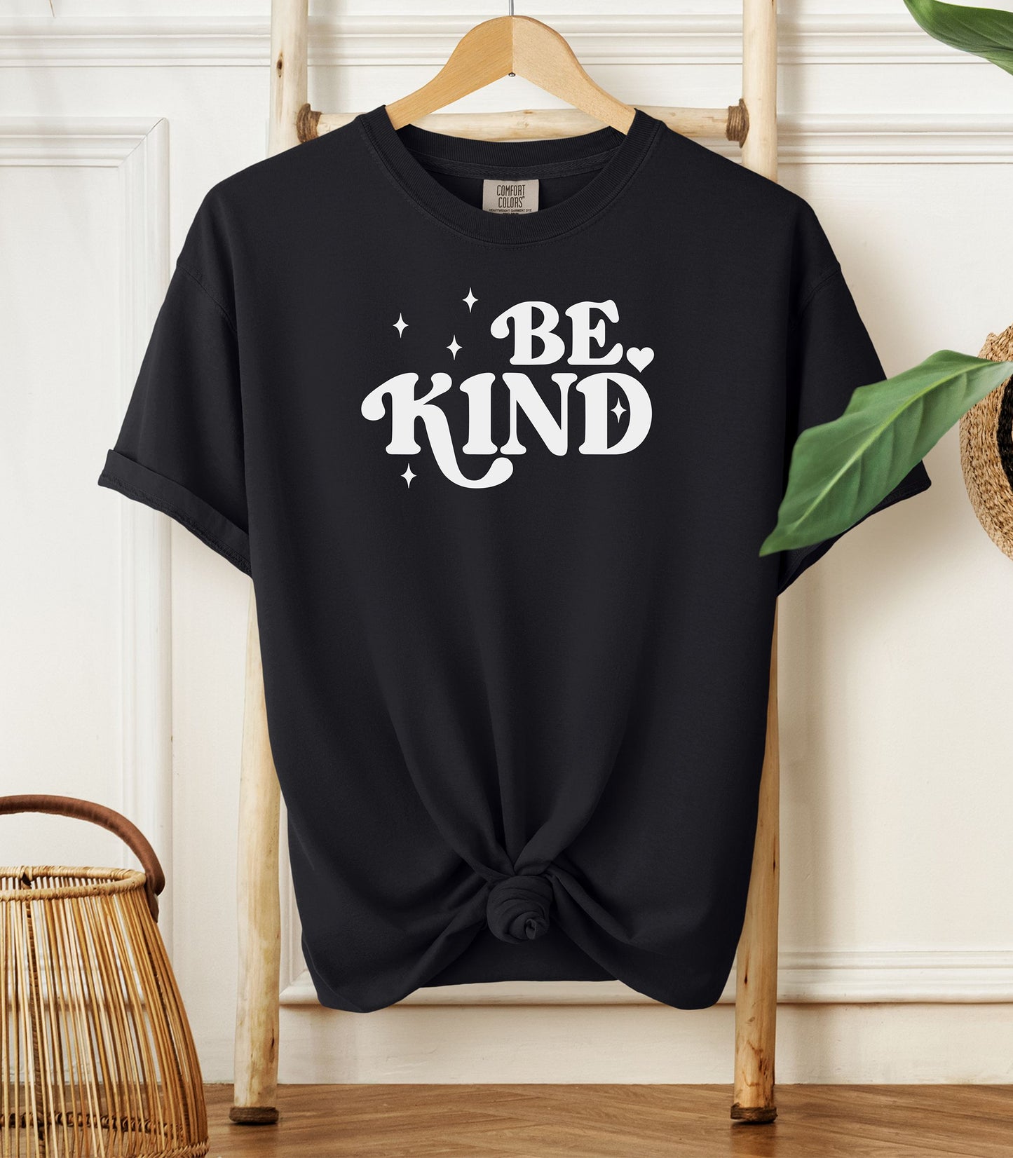 Black t-shirt with 'BE KIND' text on a wooden hanger against a white wall.
