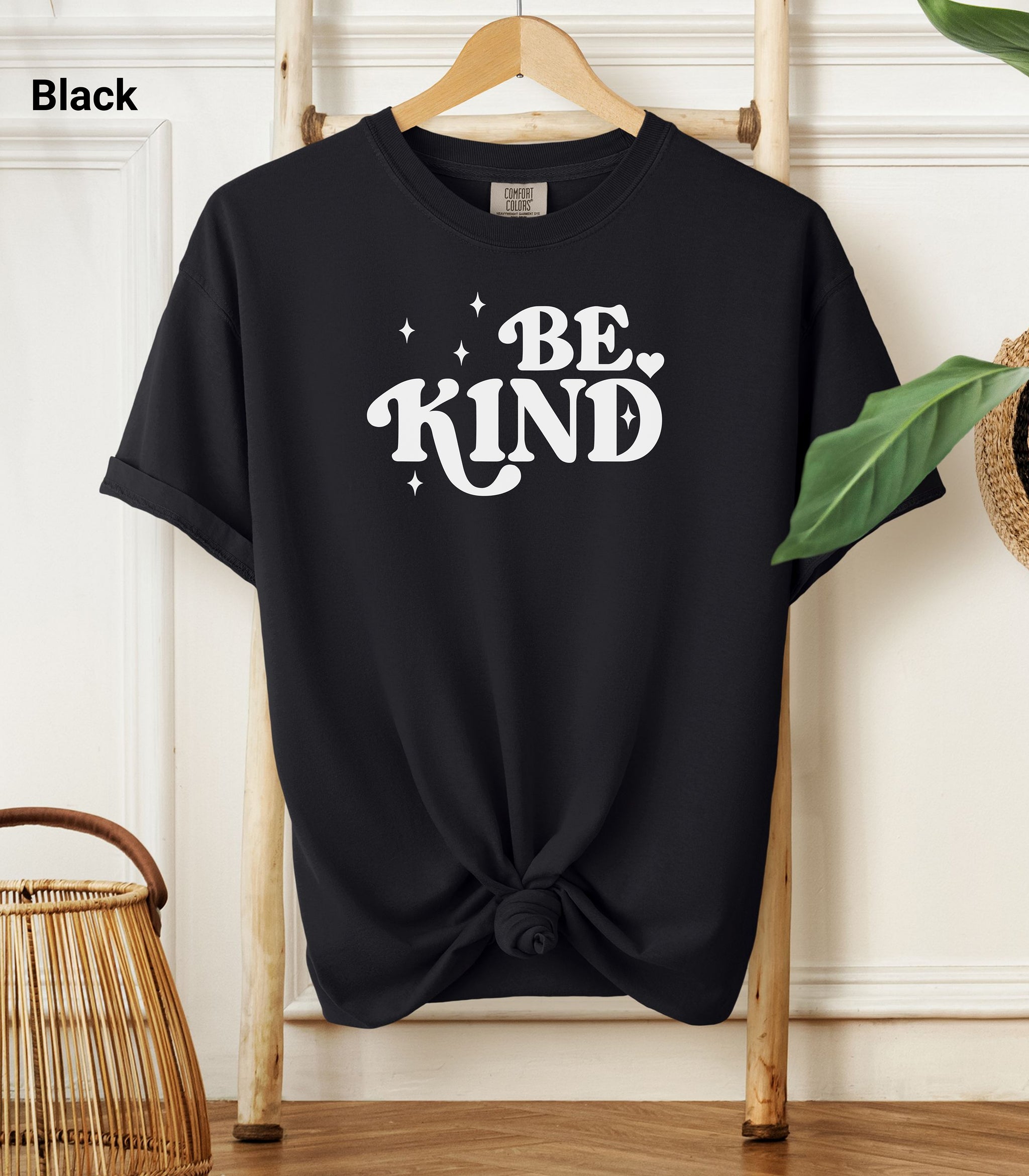Black Comfort Colors T-shirt featuring a retro-style “Be Kind” design in bold white lettering with star and heart accents.   Shirt is knotted at the bottom and displayed on a wooden hanger against a neutral background with decorative plants.