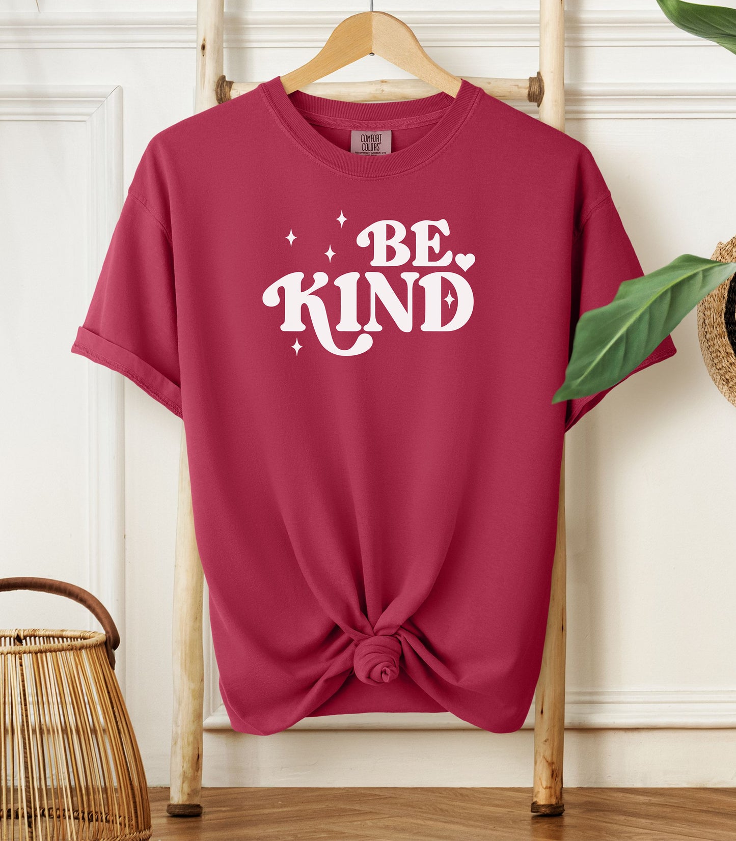 Chili t-shirt with 'BE KIND' text on a wooden hanger against a white wall.