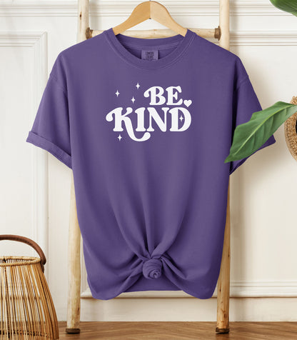 Grape purple t-shirt with 'BE KIND' text on a wooden hanger against a white wall.
