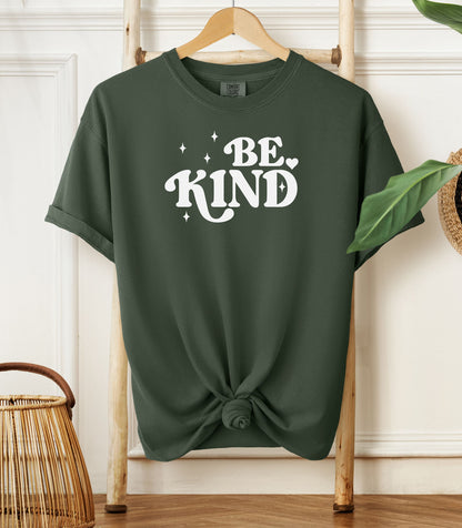 Hemp green t-shirt with 'BE KIND' text on a wooden hanger against a white wall.