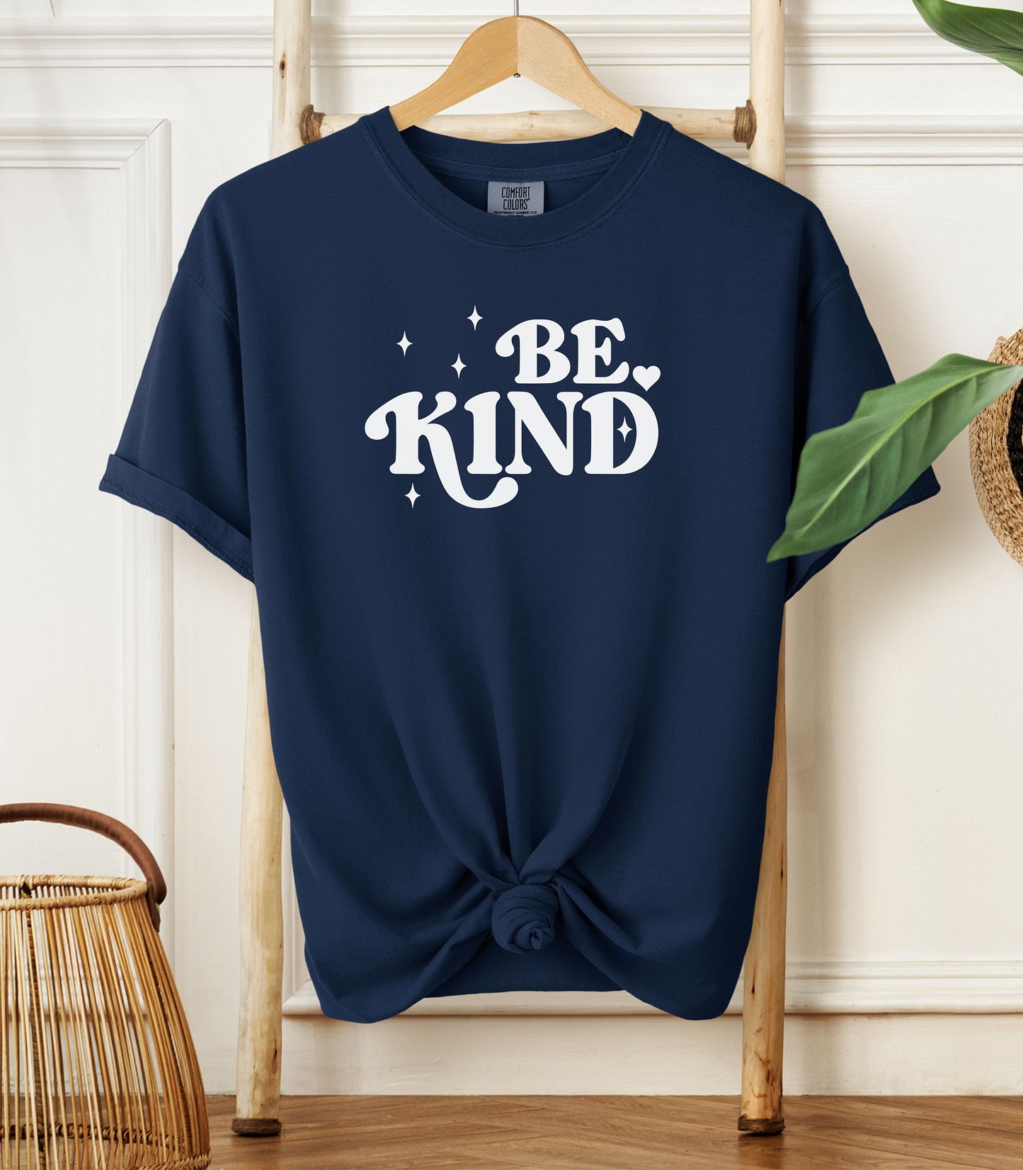 True Navy blue t-shirt with 'BE KIND' text on a wooden hanger against a white wall.