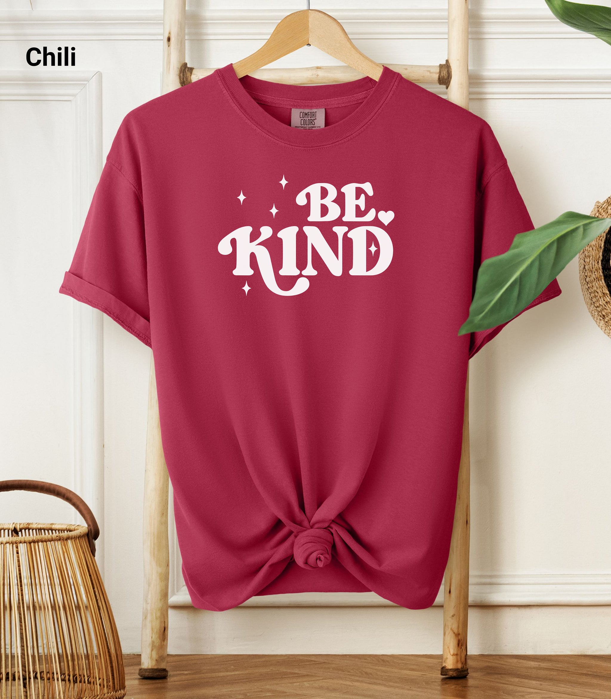 Chili color Comfort Colors T-shirt featuring a retro-style “Be Kind” design in bold white lettering with star and heart accents.   Shirt is knotted at the bottom and displayed on a wooden hanger against a neutral background with decorative plants.