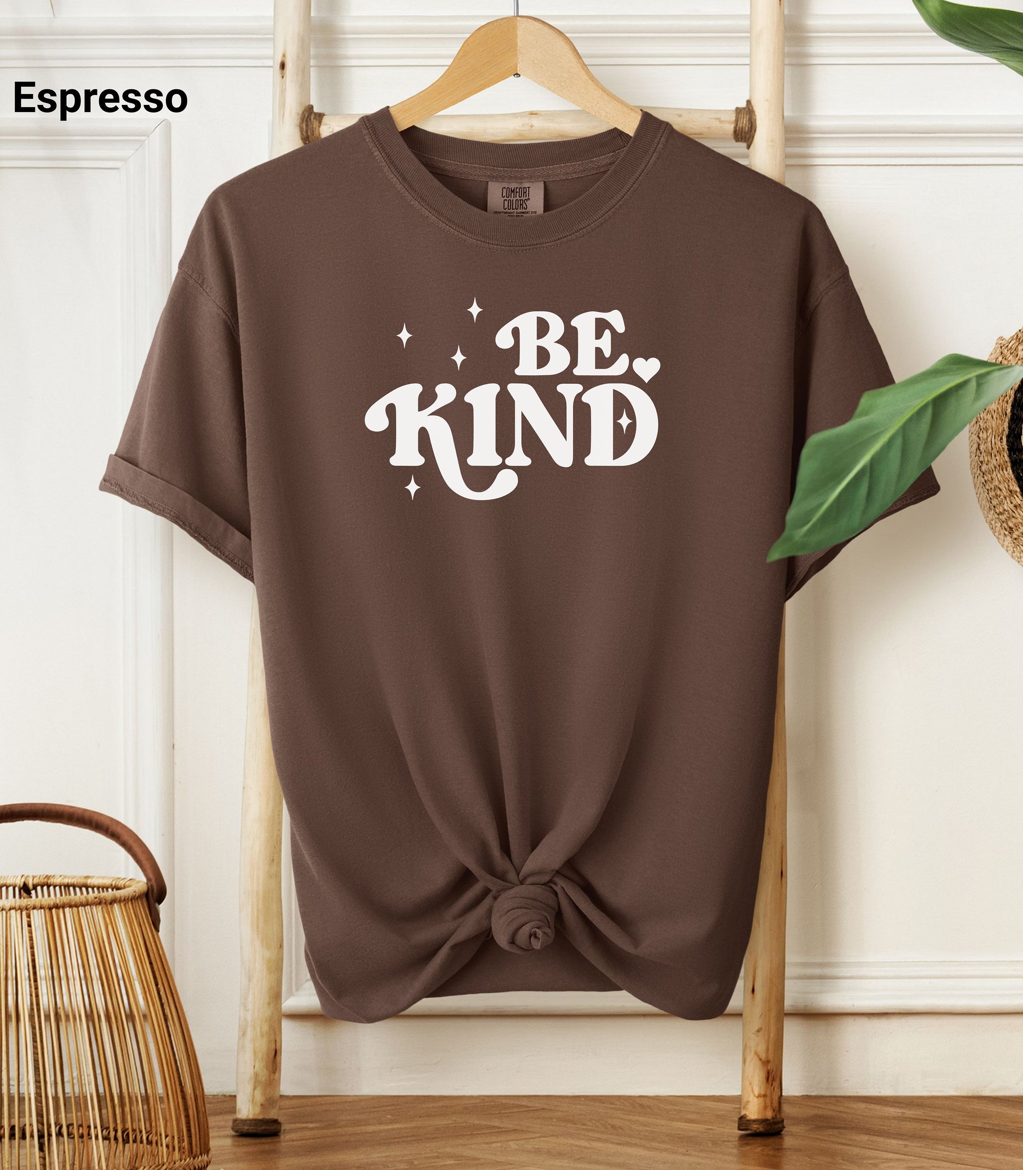  Espresso Comfort Colors T-shirt featuring a retro-style “Be Kind” design in bold white lettering with star and heart accents.   Shirt is knotted at the bottom and displayed on a wooden hanger against a neutral background with decorative plants.
