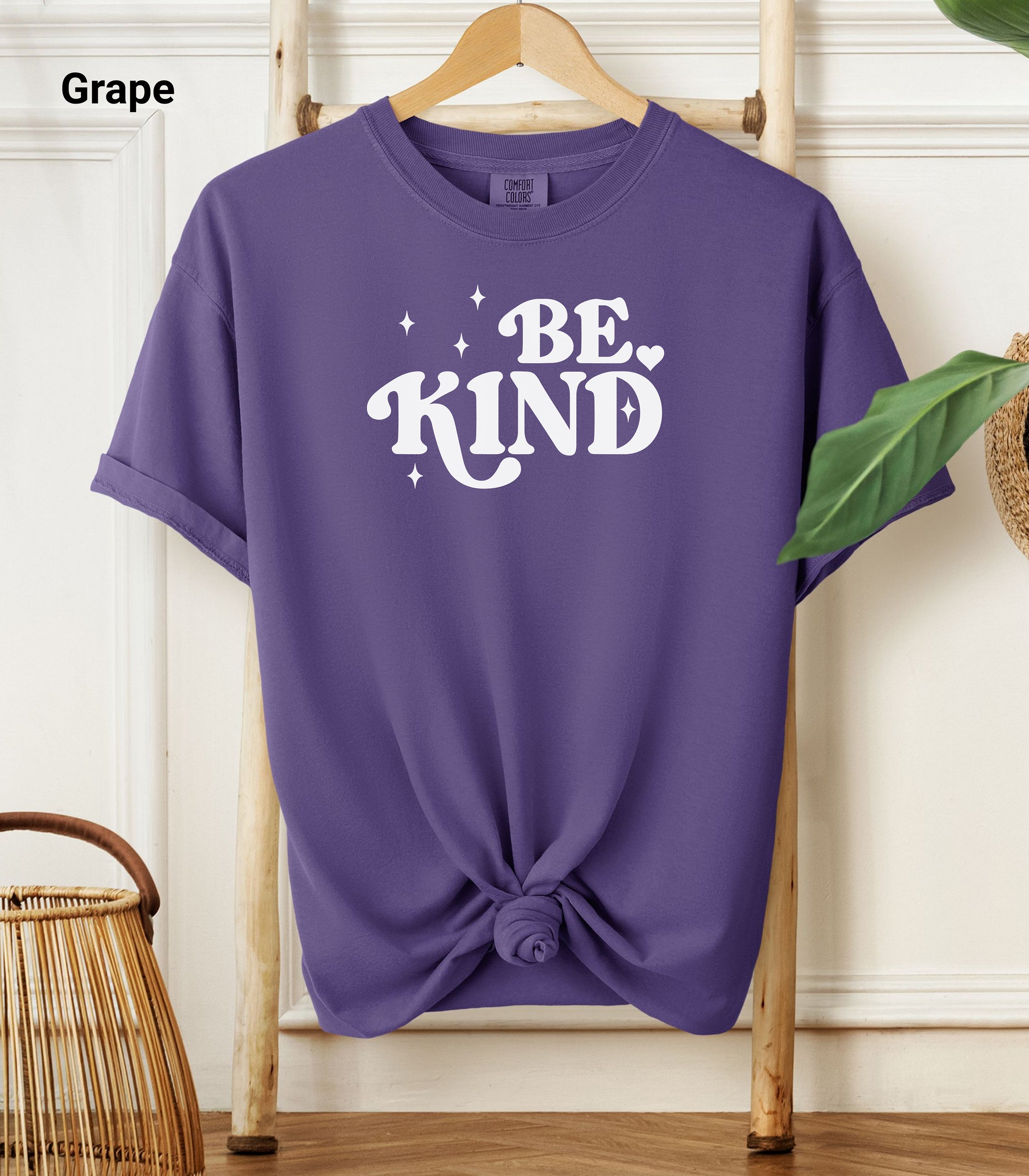 Grape Comfort Colors T-shirt featuring a retro-style “Be Kind” design in bold white lettering with star and heart accents.   Shirt is knotted at the bottom and displayed on a wooden hanger against a neutral background with decorative plants.