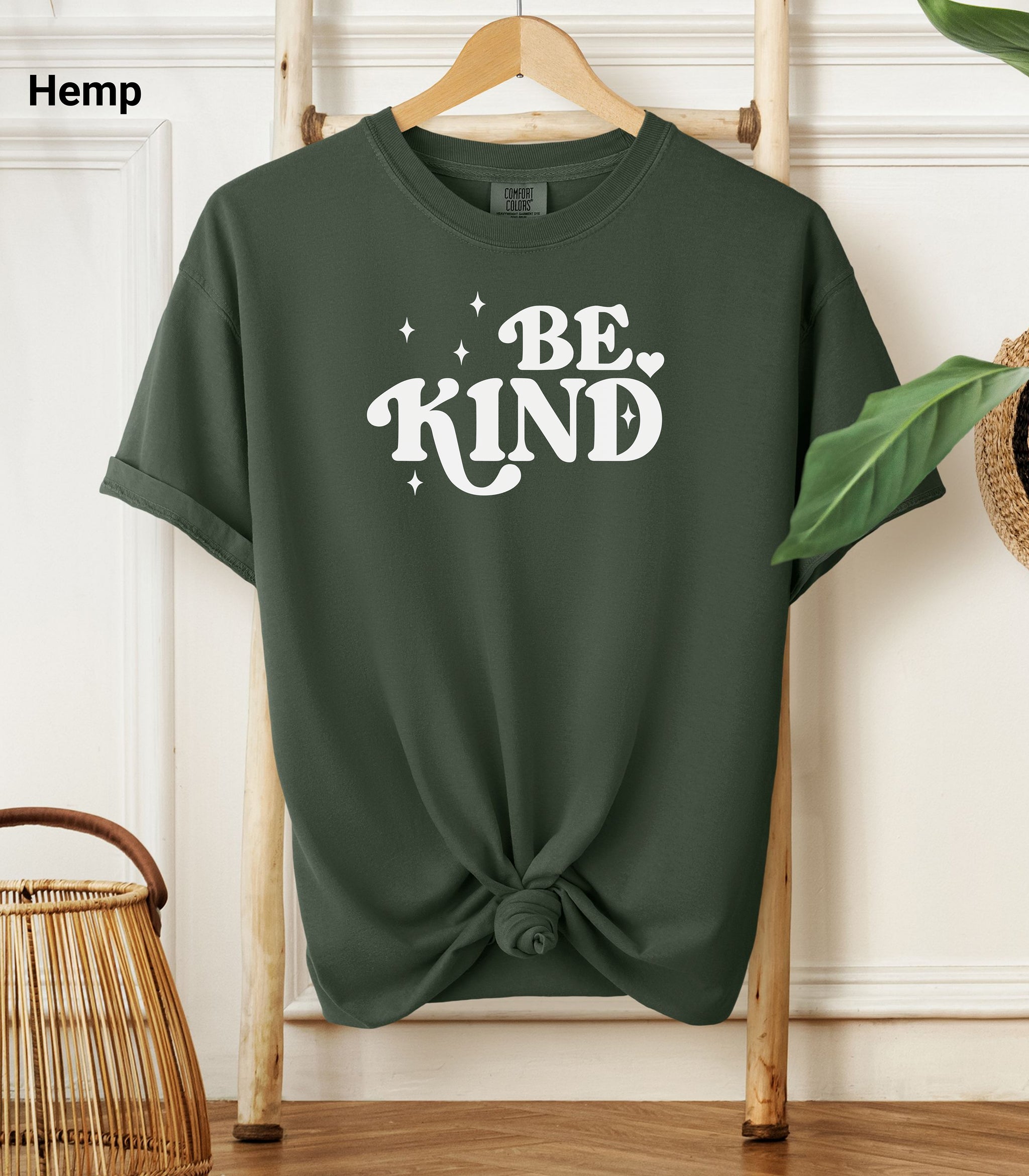 Hemp color Comfort Colors T-shirt featuring a retro-style “Be Kind” design in bold white lettering with star and heart accents.   Shirt is knotted at the bottom and displayed on a wooden hanger against a neutral background with decorative plants.