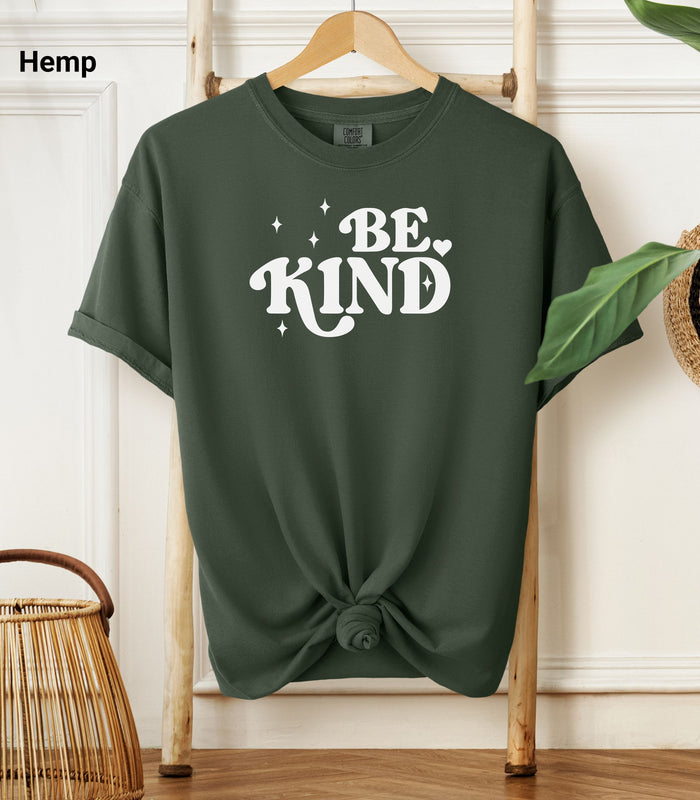 Hemp color Comfort Colors T-shirt featuring a retro-style “Be Kind” design in bold white lettering with star and heart accents.   Shirt is knotted at the bottom and displayed on a wooden hanger against a neutral background with decorative plants.