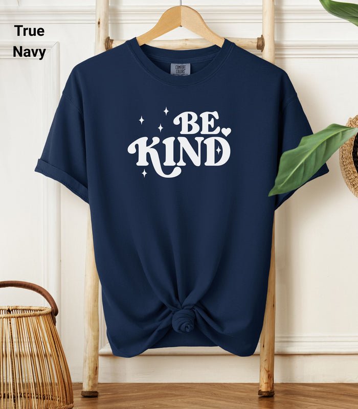True Navy Comfort Colors T-shirt featuring a retro-style “Be Kind” design in bold white lettering with star and heart accents.   Shirt is knotted at the bottom and displayed on a wooden hanger against a neutral background with decorative plants.