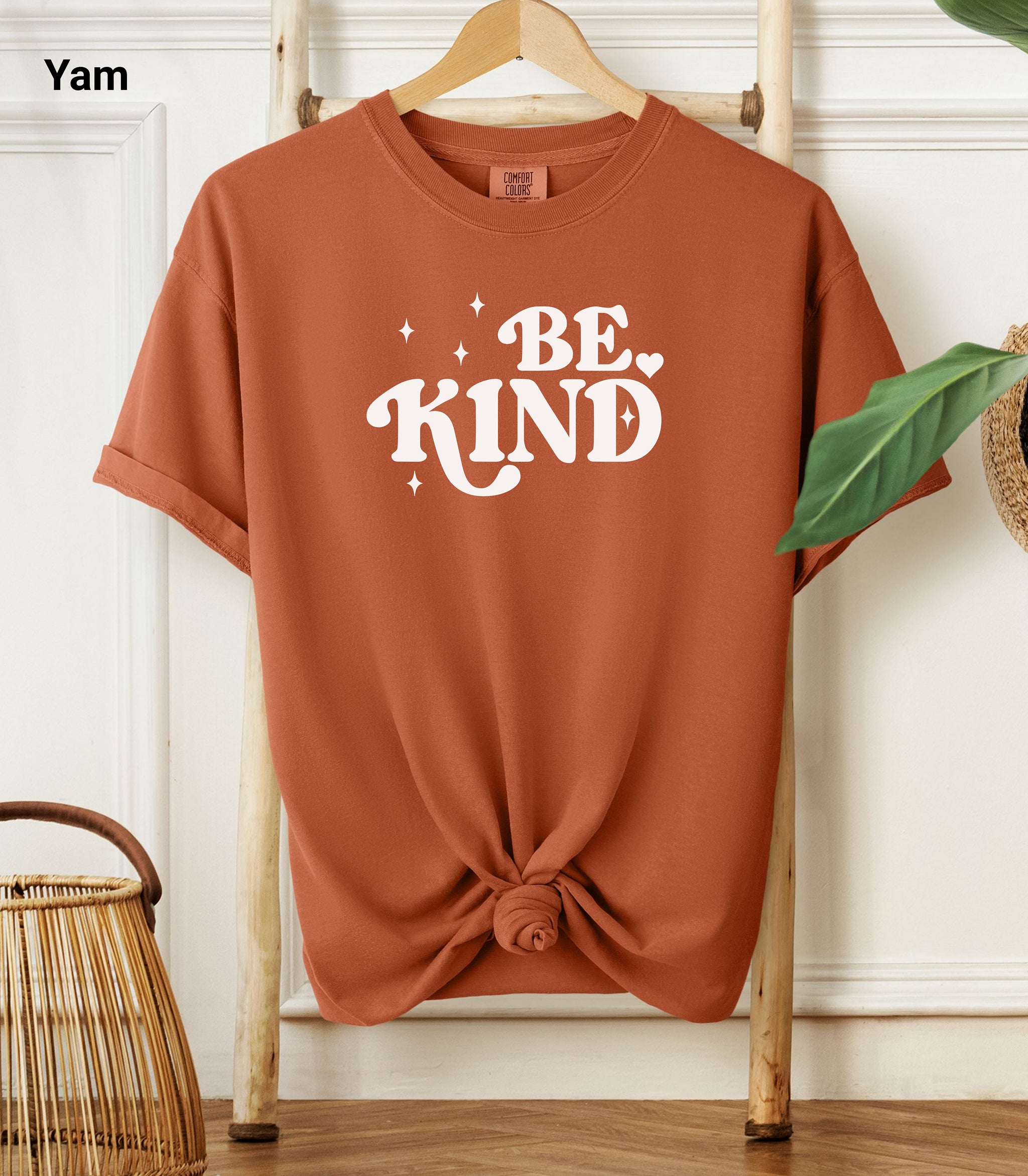 Yam Comfort Colors T-shirt featuring a retro-style “Be Kind” design in bold white lettering with star and heart accents.   Shirt is knotted at the bottom and displayed on a wooden hanger against a neutral background with decorative plants.