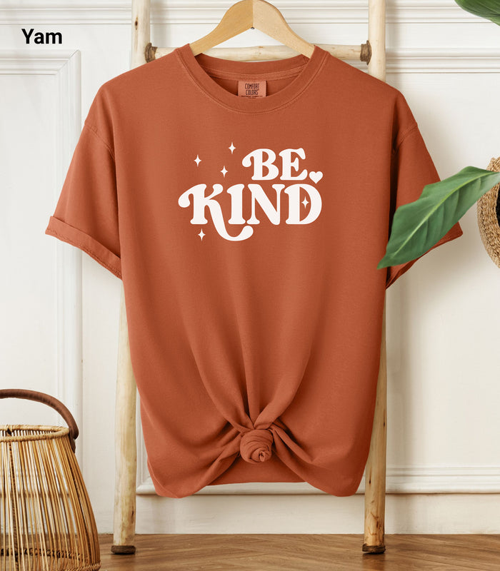 Yam Comfort Colors T-shirt featuring a retro-style “Be Kind” design in bold white lettering with star and heart accents.   Shirt is knotted at the bottom and displayed on a wooden hanger against a neutral background with decorative plants.