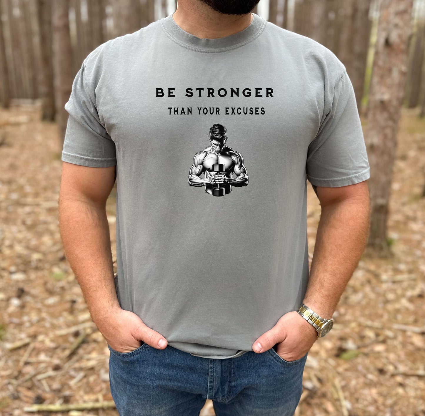 Person wearing a grey T-shirt featuring the motivational phrase “Be Stronger Than Your Excuses” in bold black lettering, with a graphic of a muscular figure holding a dumbbell below the text. Perfect for gym-goers, fitness enthusiasts, and athletes, this empowering workout shirt inspires determination, discipline, and strength training focus.